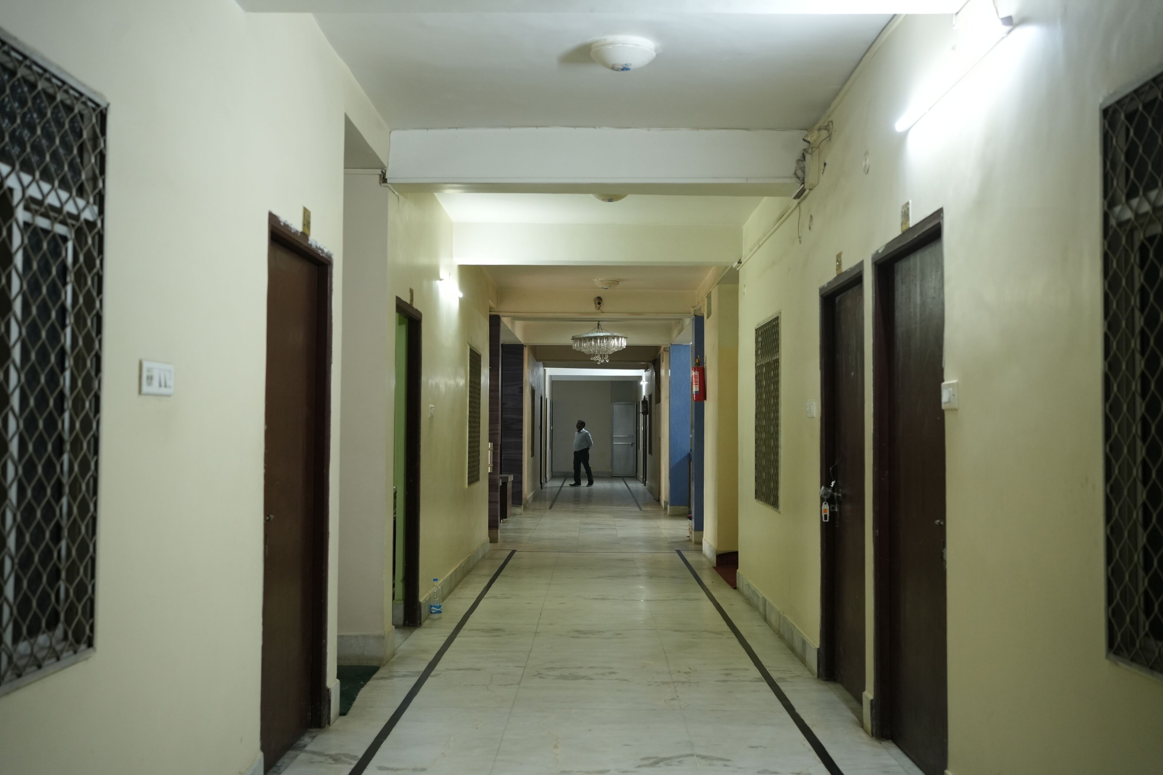 A corridor with doors on both sides, a person walking down the hallway, and a chandelier hanging from the ceiling.