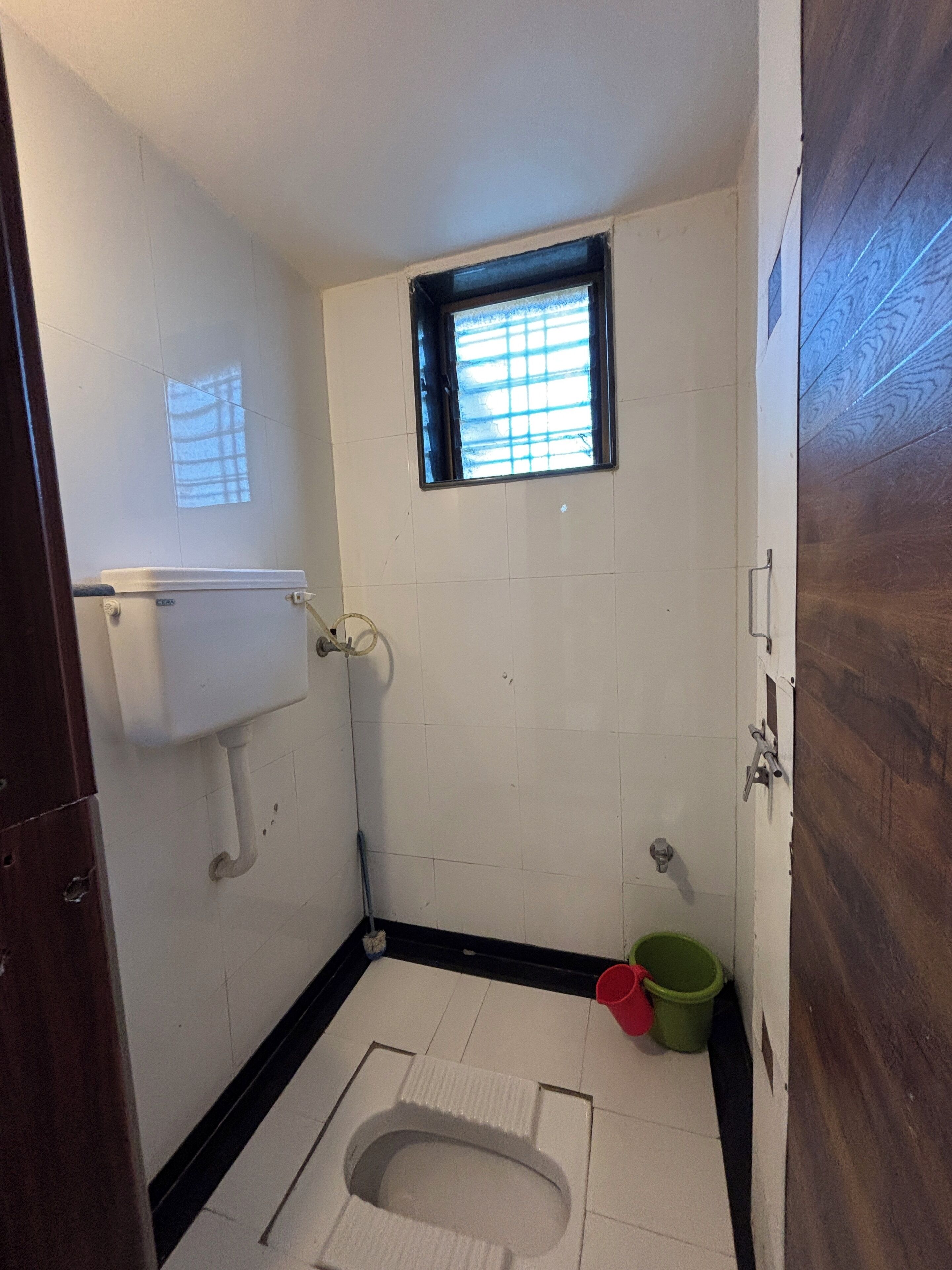 Un baño pequeño y estrecho con paredes de azulejos blancos, una ventana con persianas, un inodoro, un lavamanos y un cubo verde.