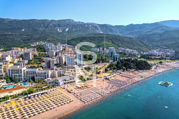 Paesaggio costiero con spiaggia, alberghi e montagne sullo sfondo.