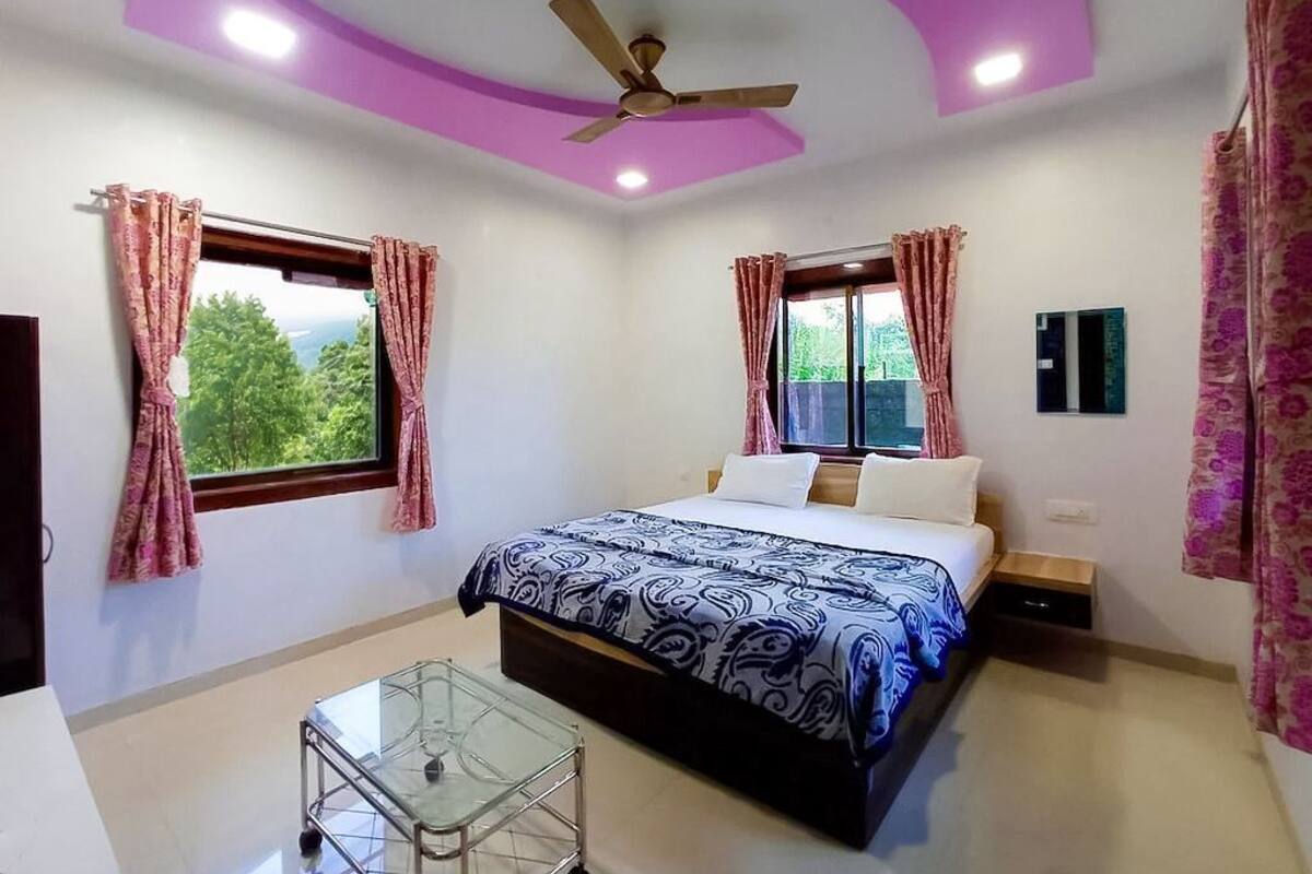 A bedroom with a bed, ceiling fan, two windows with curtains, a bedside table, and a glass table.