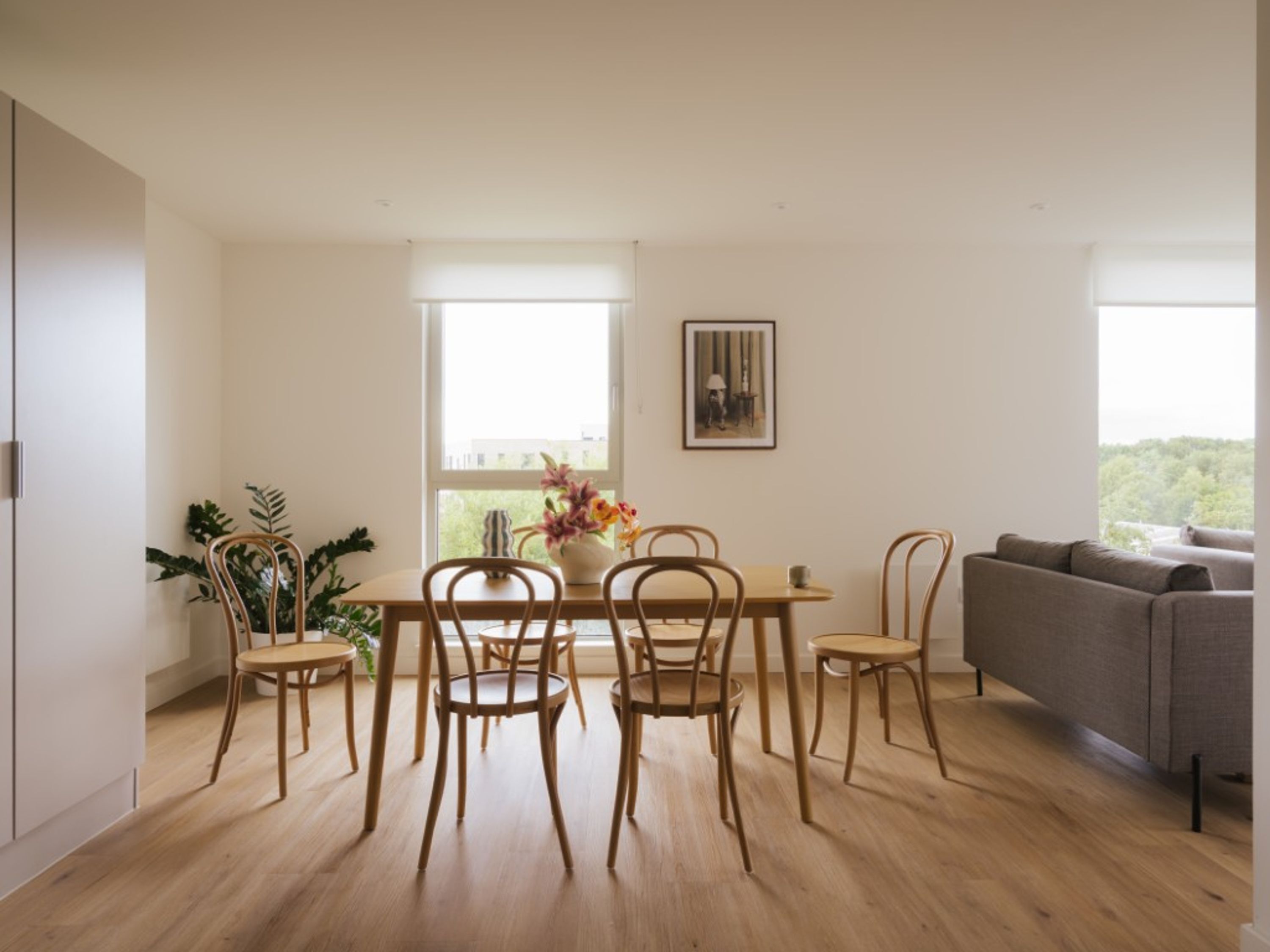 A modern living room with a dining area, featuring a wooden table and chairs, a grey sofa, and a large window with a view.