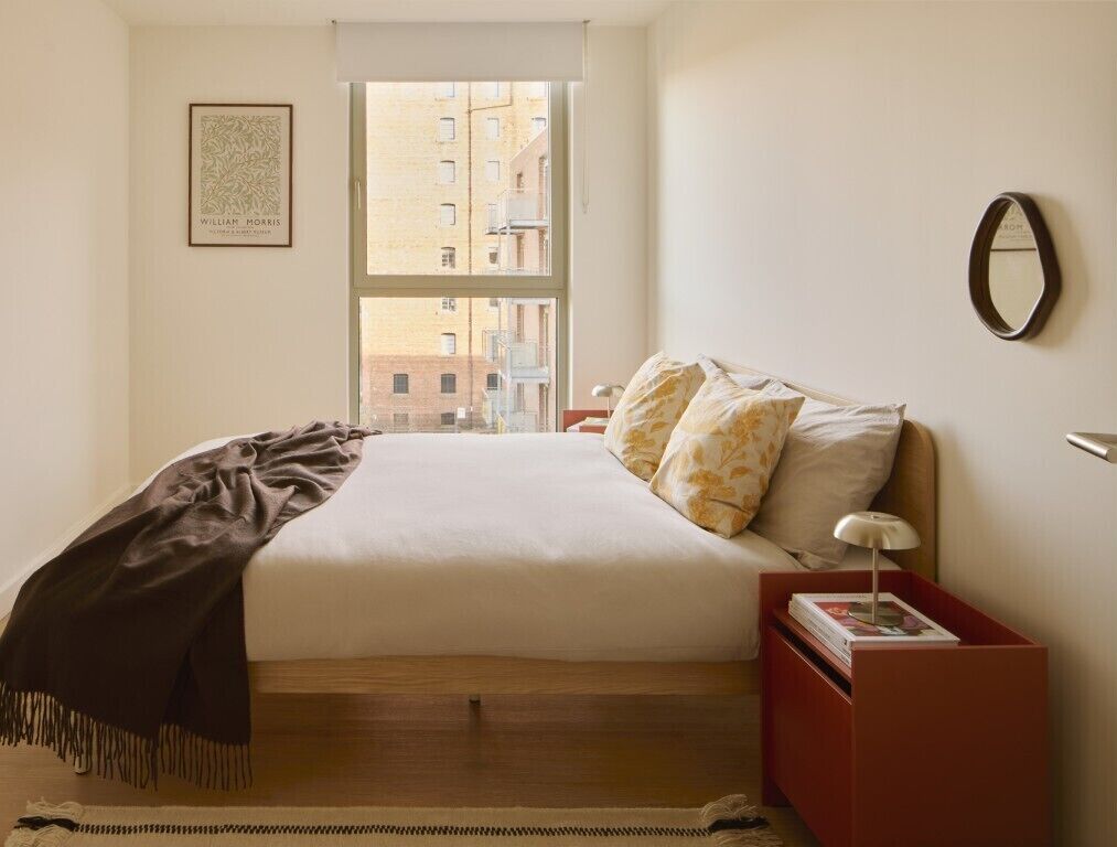 A neatly arranged bedroom with a bed, a bedside table, a nightstand, a window, and a framed picture on the wall.