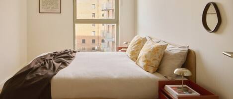 A neatly arranged bedroom with a bed, a bedside table, a nightstand, a window, and a framed picture on the wall.