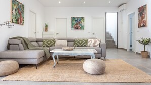 Ein modernes Wohnzimmer mit einem grauen Ecksofa, einem weißen Couchtisch und einem großen Webteppich. Zwei Türen führen in andere Räume, im Hintergrund ist eine Treppe zu sehen.