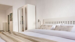 Ein ordentlich gemachtes Bett mit weißem Kopfteil, einer beige farbenen Tagesdecke und einem weißen Kleiderschrank mit Spiegel.