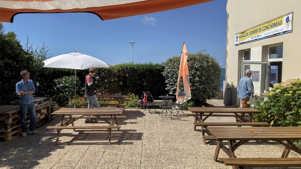 Un espace extérieur aménagé en terrasse, avec des tables de pique-nique en bois et des parasols, un bâtiment avec une enseigne, et de la verdure en arrière-plan.
