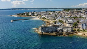 Vue aérienne d’une ville côtière, avec un imposant ensemble de bâtiments situé près du front de mer, une jetée s’avançant dans la mer et une plage de sable.