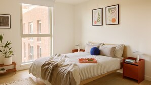 Een slaapkamer met een groot raam, een bed met een gestreept dekbedovertrek, twee aan de muur bevestigde fotolijstjes, een nachtkastje met een lamp en een kleine potplant.