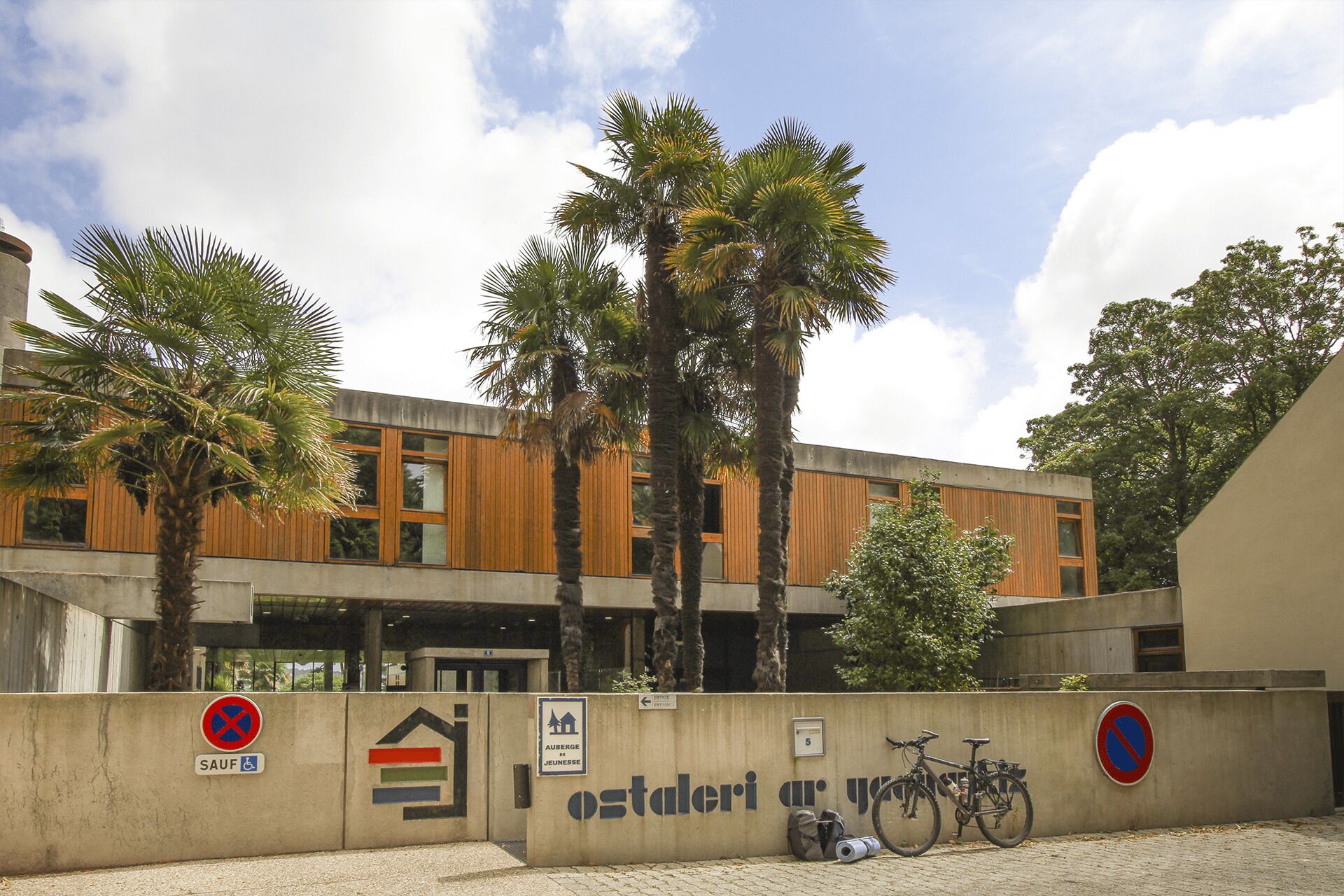 Un bâtiment moderne avec une façade en béton, un revêtement mural en bois et des palmiers. On y trouve des panneaux indiquant les restrictions de stationnement et le symbole de stationnement réservé aux personnes handicapées.