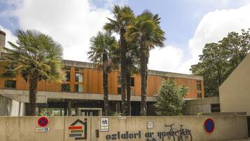 Un edificio moderno con facciata in cemento, rivestimenti in legno e palme. Sono presenti segnali che indicano divieti di parcheggio e il simbolo per posti auto riservati ai disabili.