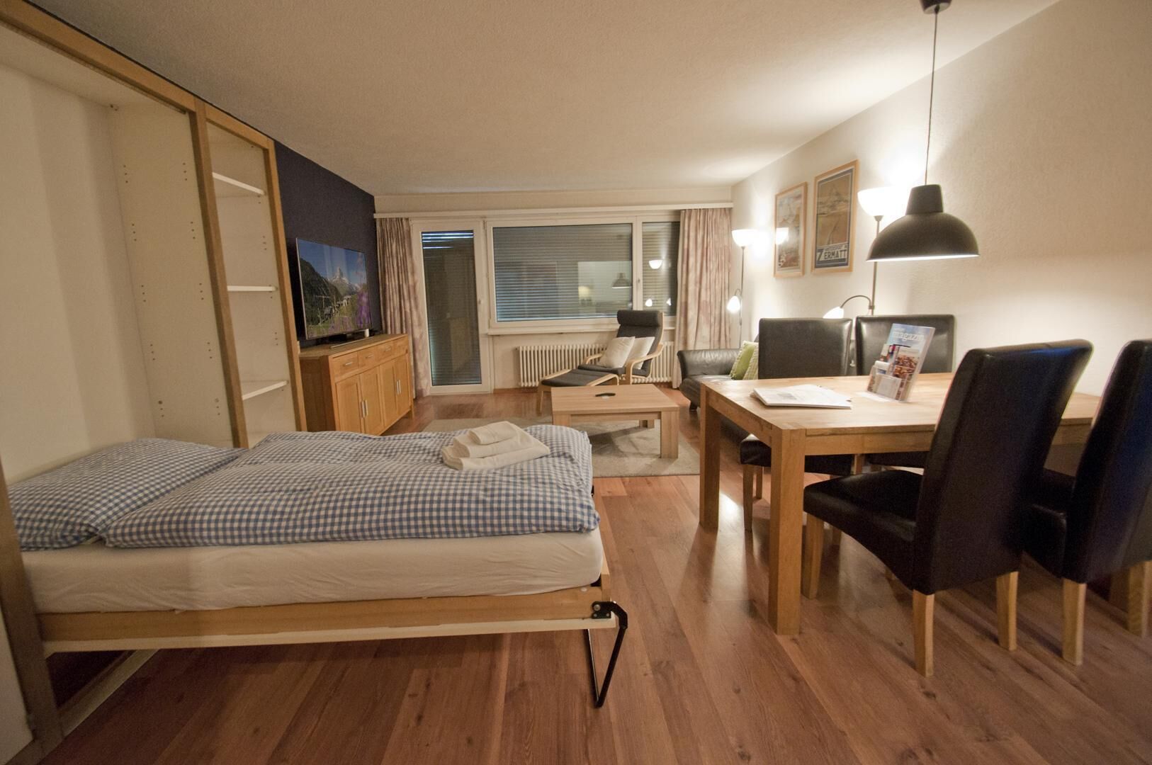 A compact room with a bed, a desk, two chairs, a TV, and a window with curtains.