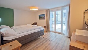 A hotel room with a double bed, a wooden floor, a small wooden stool, a bathroom with a sink, and a large window with a view of a building and snow outside. - Chalet Aeschhorn, Apartment Lucy (Zermatt)