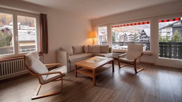 Ein modernes Wohnzimmer mit einer Couch, zwei Sesseln, einem Couchtisch und einer Lampe. Große Fenster bieten Blick auf eine Berglandschaft.