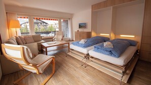 Ein modernes Hotelzimmer mit einer Couch, zwei Betten, einem kleinen Tisch und Blick auf die Stadt durch große Fenster.