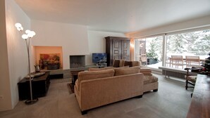 Una sala de estar moderna con un sofá, un televisor de pantalla plana, una chimenea y un ventanal que ofrece vistas al exterior.