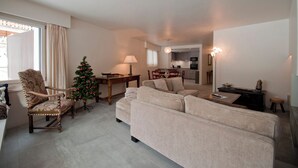 Una sala de estar con un sofá color beige, una silla de madera, un pequeño árbol de Navidad y un comedor al fondo.