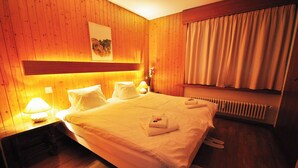 Habitación de hotel con paneles de madera, una cama con sábanas blancas, una mesita de noche con lámpara y un radiador.