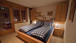 Een slaapkamer met een houten bed, geruit beddengoed, een nachtkastje met lamp, een raam met zonwering en houten planken met boeken en een teddybeer.