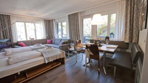 A hotel room with a bed, sofa, dining table, and chairs. There are curtains, a window, and a lamp. - Haus Bodmen C, Apartment Edelweiss (Zermatt)