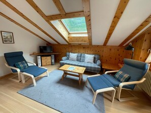 Una habitación acogedora con techo inclinado, vigas de madera, un sofá, dos sillones, una mesita y un televisor.