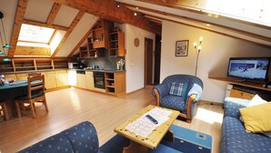 Ein gemütlicher, hell erleuchteter Raum mit Esstisch, blau gemustertem Sofa und einem Fernseher, der einen Online-Ticketshop anzeigt.