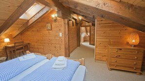 Um quarto de sótão acolhedor, com paredes de madeira, teto inclinado e um claraboia. Há duas camas com roupa de cama xadrez e um aparador de madeira com uma lâmpada.