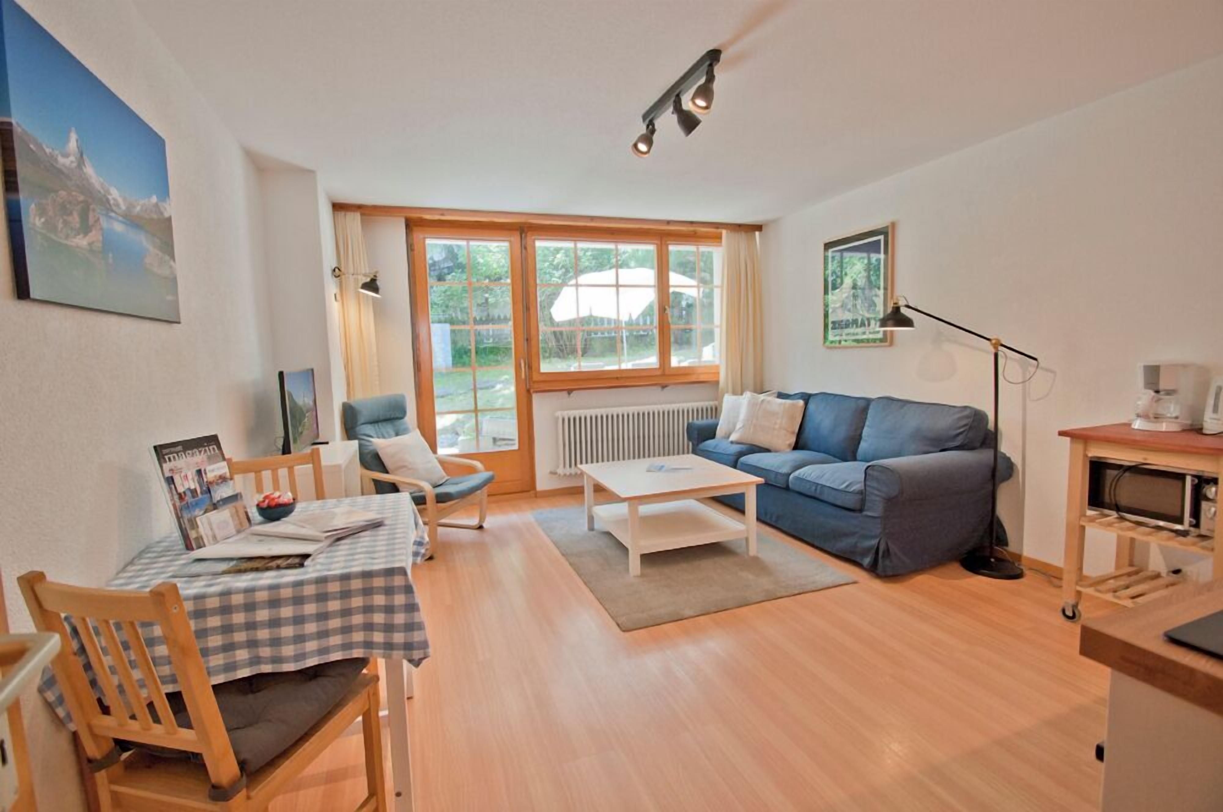 Ein Wohnzimmer mit einem blauen Sofa, einem Esstisch mit kariertem Tischläufer, Holzstühlen, einem weißen Couchtisch und einem großen Fenster mit Vorhängen.