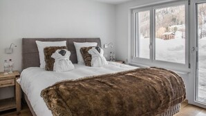 A bedroom with a large bed, two white pillows, a brown fur throw, and a view of a snowy landscape through a window. - Haus Andy, Apartment Andy (Zermatt)