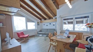 Ein modernes Interieur mit Holzbalkendecke, einer Essecke mit Tisch und Stühlen, einem roten Sessel, einer Couch und einem Fenster mit Blick.