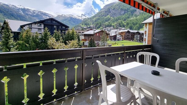A balcony with a table and chairs, overlooking a mountainous landscape with houses and trees. - Haus Matten, Studio Findeln (Zermatt)
