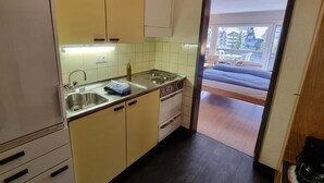 A compact kitchen with a sink, stove, and cabinets. A bed is visible through an open door, leading to a room with a view of buildings. - Haus Matten, Studio Findeln (Zermatt)