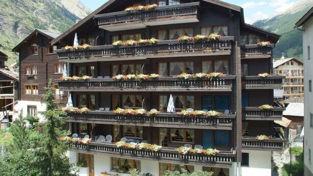 Ein mehrstöckiges Hotel mit Balkonen, die mit Blumen geschmückt sind, vor einer Bergkulisse.