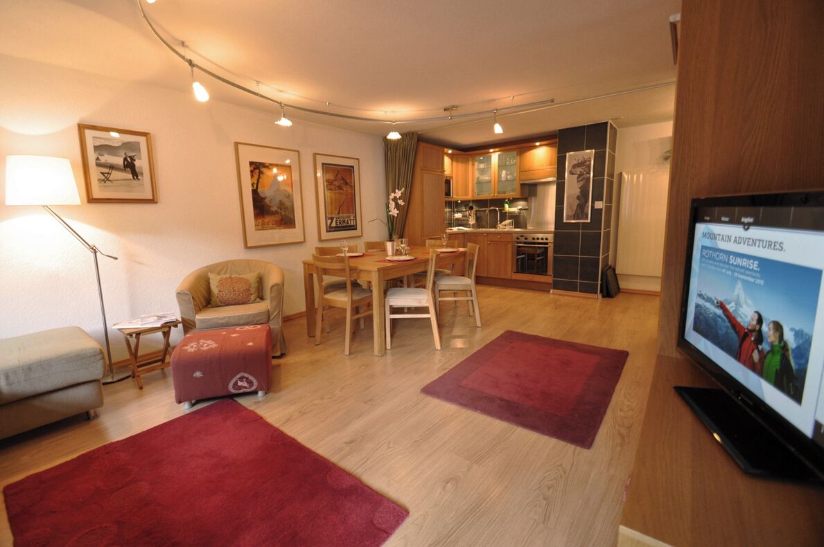 A compact living space with a dining area, kitchenette, and a television displaying 'Mountain Adventures.'