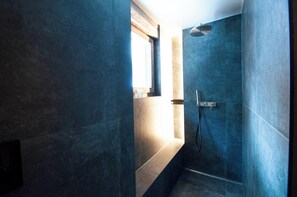Un baño moderno con azulejos azul oscuro, una ducha con cabezal de ducha manual y un ventanal que proporciona luz natural.
