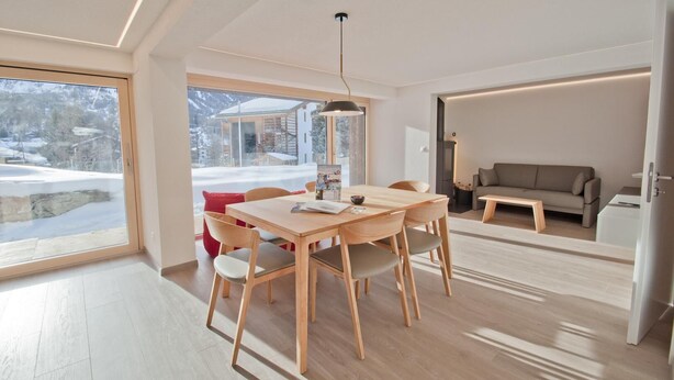 Uma sala de estar moderna com uma mesa de jantar de madeira, um sofá e uma janela grande que oferece vista para uma paisagem nevada.