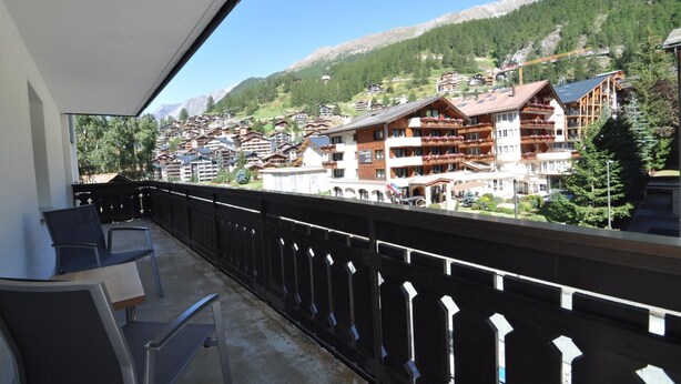 Uma varanda com vista para um resort de montanha, com vários edifícios, incluindo um hotel com varandas e um teleférico ao fundo.