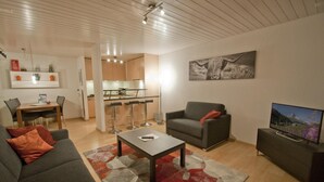 A modern living room with a sofa, a coffee table, and a TV. There is a kitchen area with a dining table and chairs. - Haus Arola, Apartment Hirle (Zermatt)