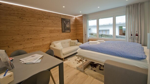 A modern hotel room with a wooden wall, a bed with a checkered duvet, a sofa, a desk with papers, and a window with a view of buildings. - Haus Aosta, Studio Aosta (Zermatt)