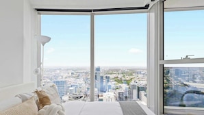 A modern bedroom with a large window overlooking a cityscape, featuring a bed with white linens and decorative pillows. - Cozy Apt Next to Southern Cross With Amazing Views (Melbourne)
