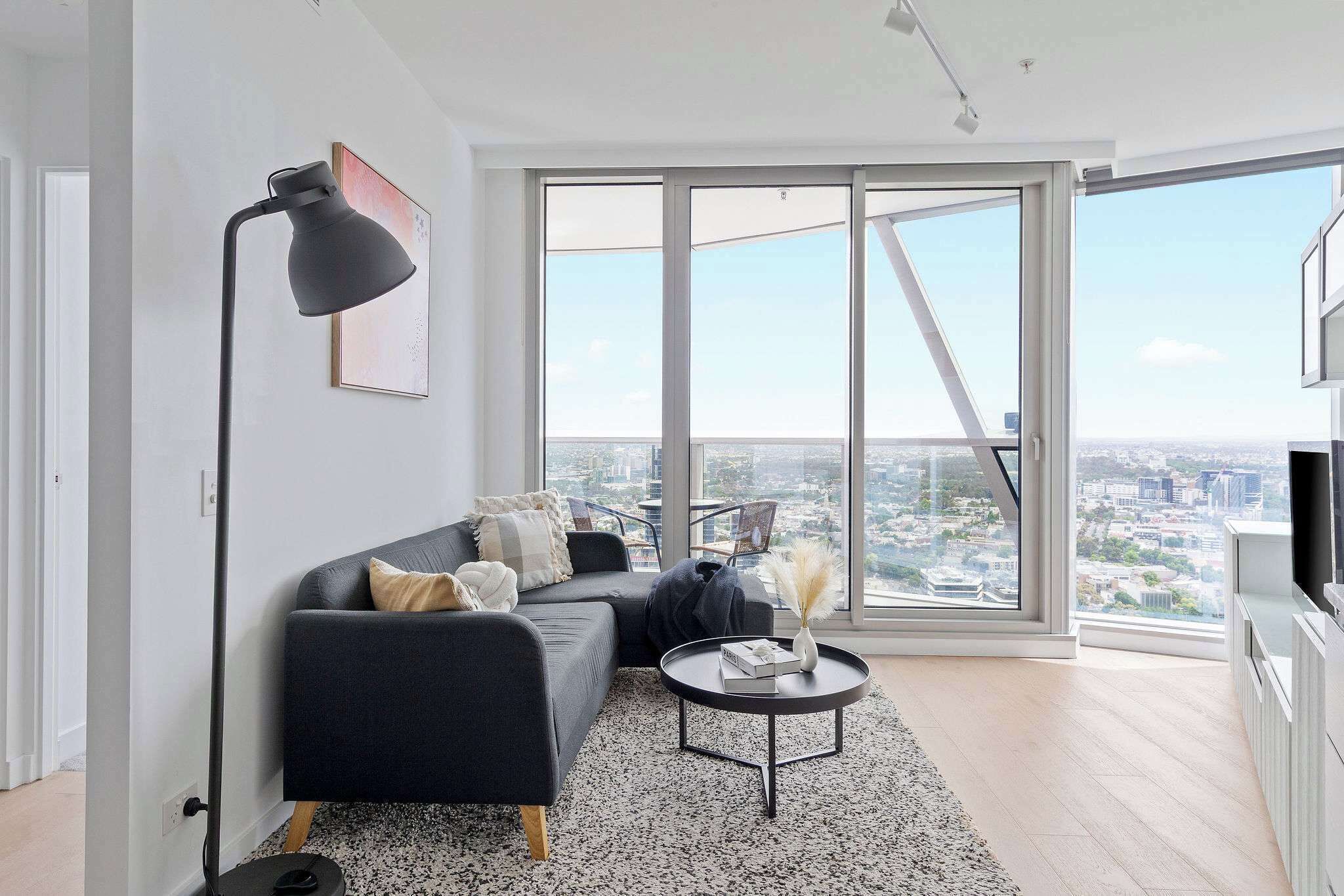 Un salon moderne avec un canapé d’angle, une table basse ronde et un lampadaire. De grandes fenêtres offrent une vue sur la ville.