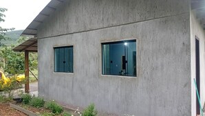 Un bâtiment en béton avec deux fenêtres, un toit en métal et un espace jardin agrémenté de plantes et d’un véhicule jaune.