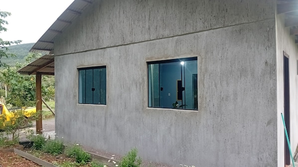 Edificio de hormigón con dos ventanas, techo metálico y un jardín con plantas y un vehículo amarillo.