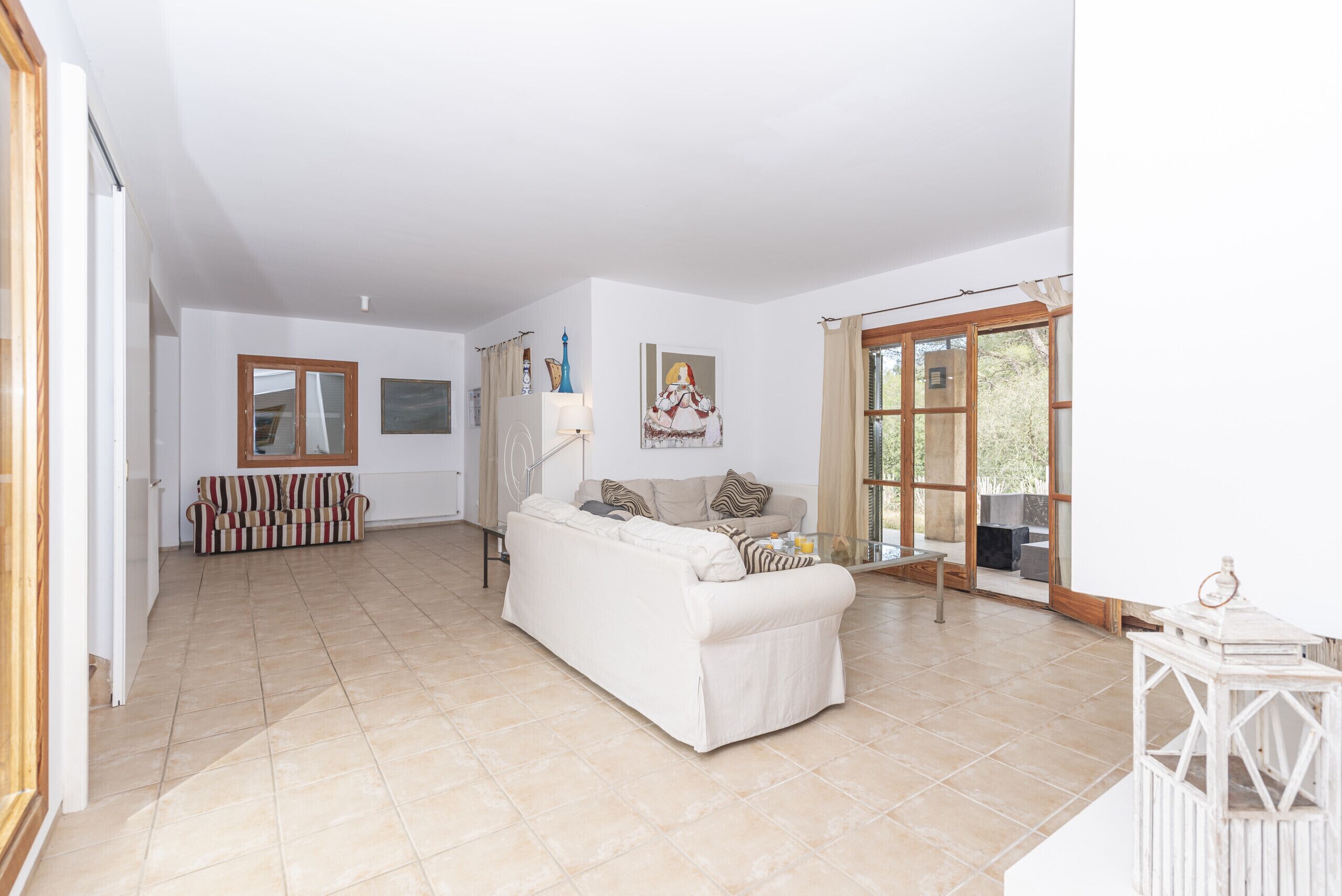 A spacious living area with a white sofa, a striped sofa, and a glass coffee table. There is a large window with curtains and a view of greenery outside.