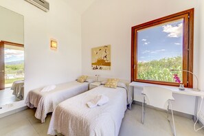 A hotel room with two single beds, a desk, a chair, a large window with a view of greenery, and a painting on the wall. - Beautiful villa with mountain views, 5km to beach (Alcúdia)