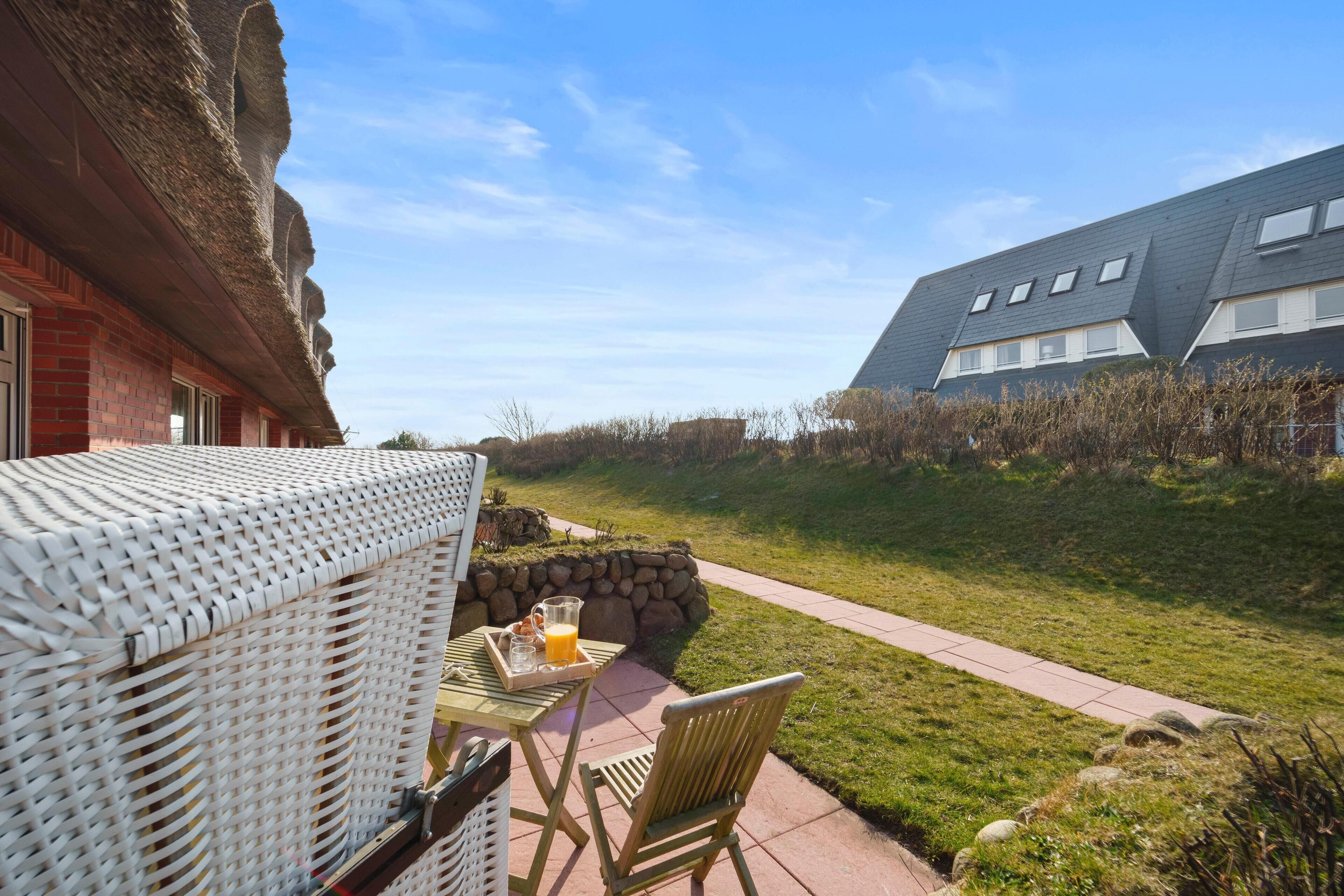 A patio with wicker furniture and a table set for breakfast, overlooking a grassy area and a building with a sloped roof.