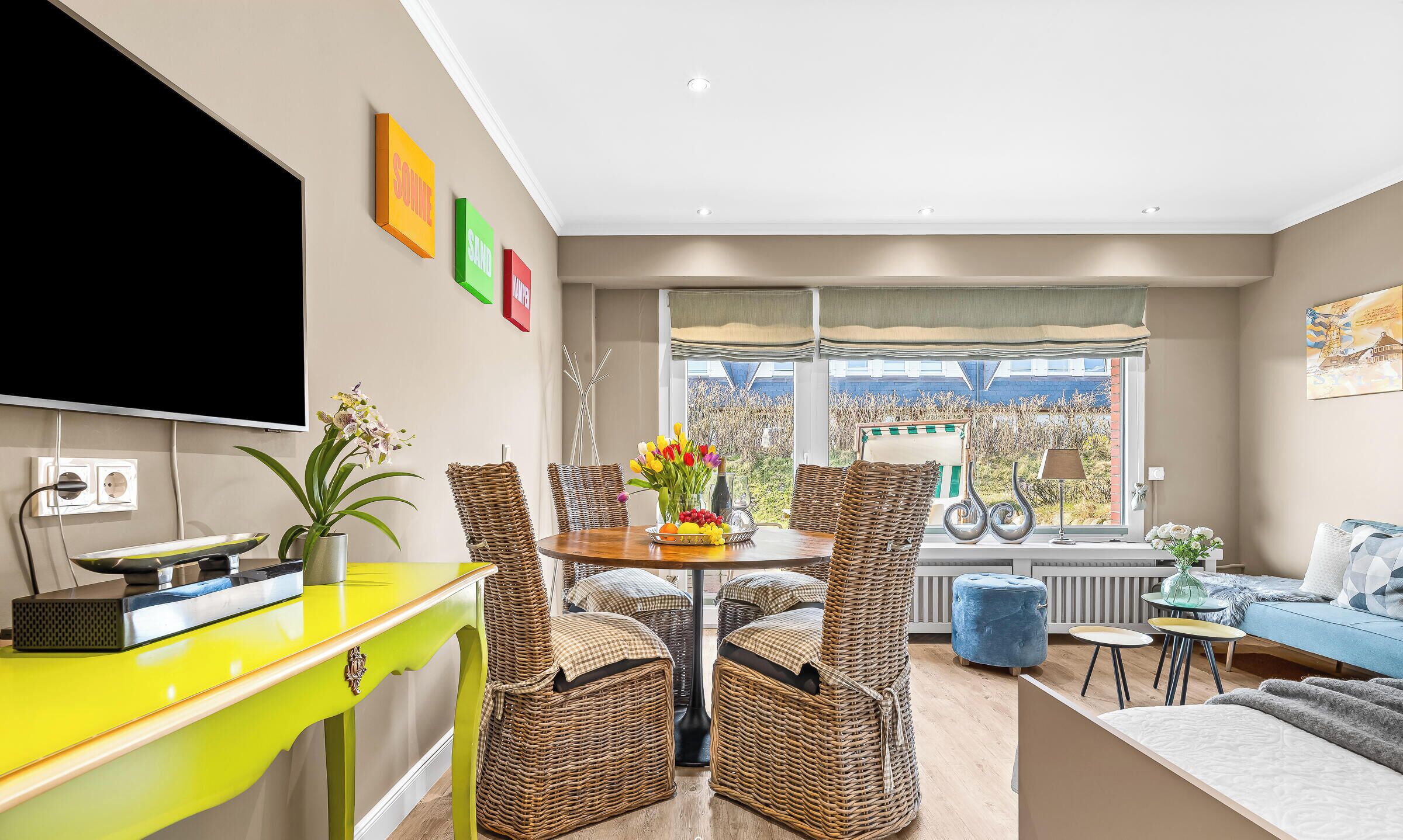 A modern living room with a dining area, wicker furniture, a bright yellow table, and a large window with a view.