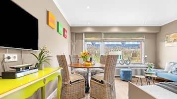A modern living room with a dining area, wicker furniture, a bright yellow table, and a large window with a view.