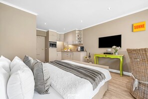 Un dormitorio moderno con cama, televisión, una kitchenette pequeña y una silla de mimbre.