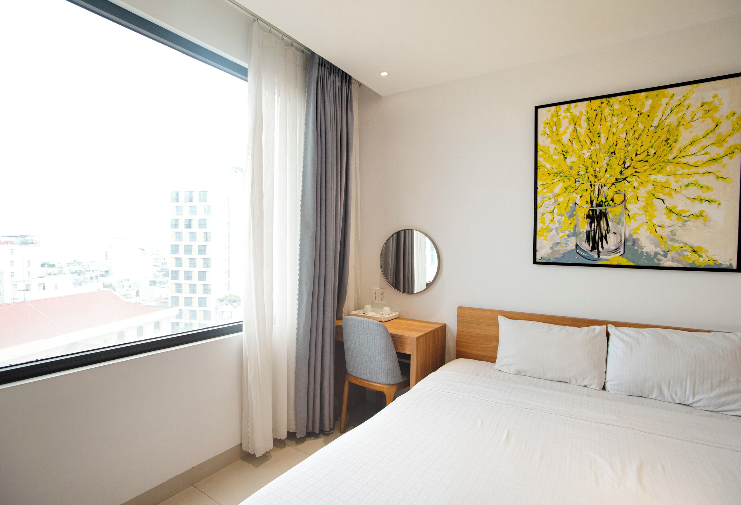 A hotel room with a bed, a desk, a chair, a round mirror, and a painting on the wall.