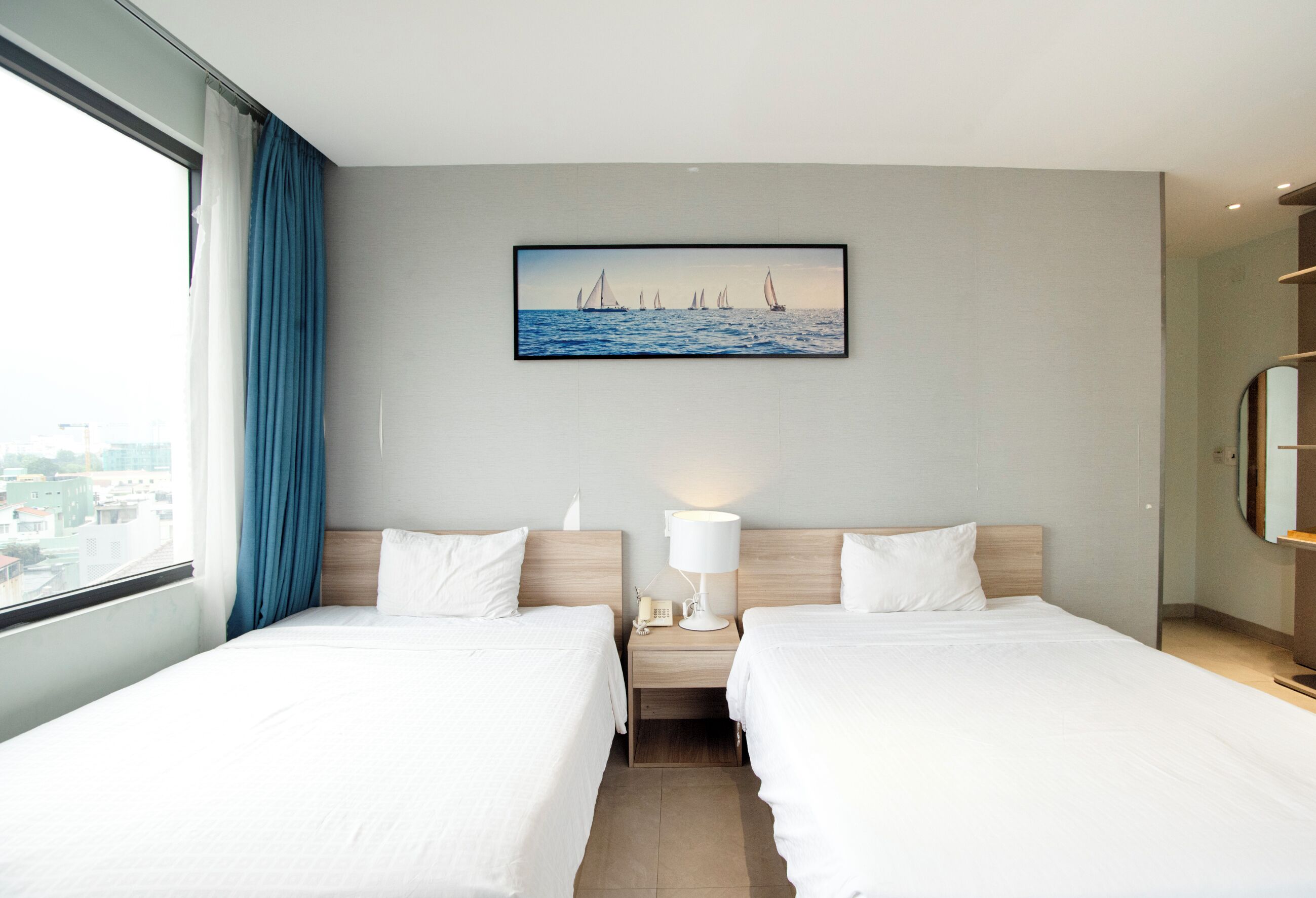 A hotel room with two beds, a nightstand with a lamp, a painting of sailboats, and a view of the city through the window.